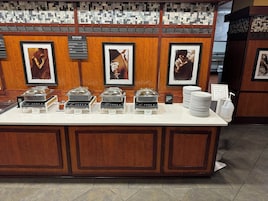 Zona de buffet con diversas estaciones de comida, incluyendo una sección de alimentos calientes, y un cartel que indica que el buffet de desayuno cuesta $14.95 más impuestos.