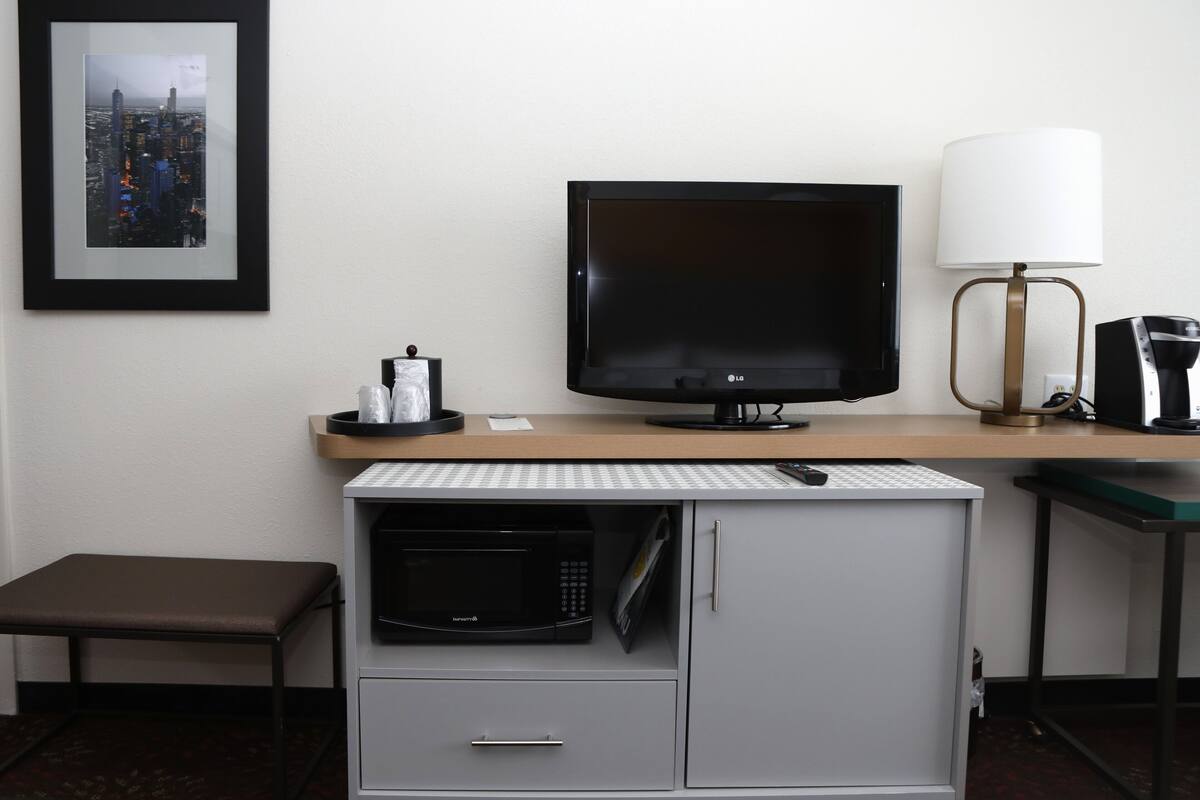 A hotel room with a flat-screen TV, a microwave, a small table with a lamp, and a framed picture on the wall.