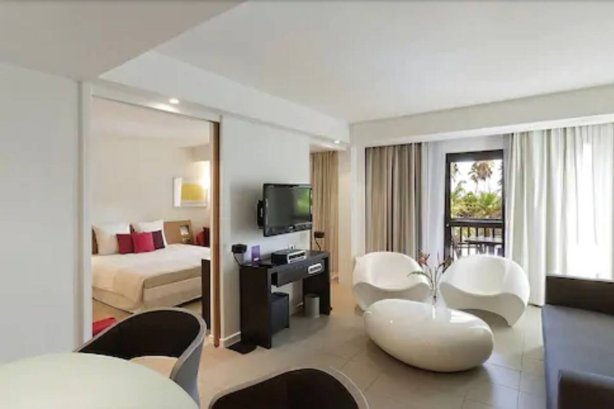 Une chambre d’hôtel comprenant un lit, un bureau avec une télévision, un coin salon avec des fauteuils et un balcon avec des rideaux.