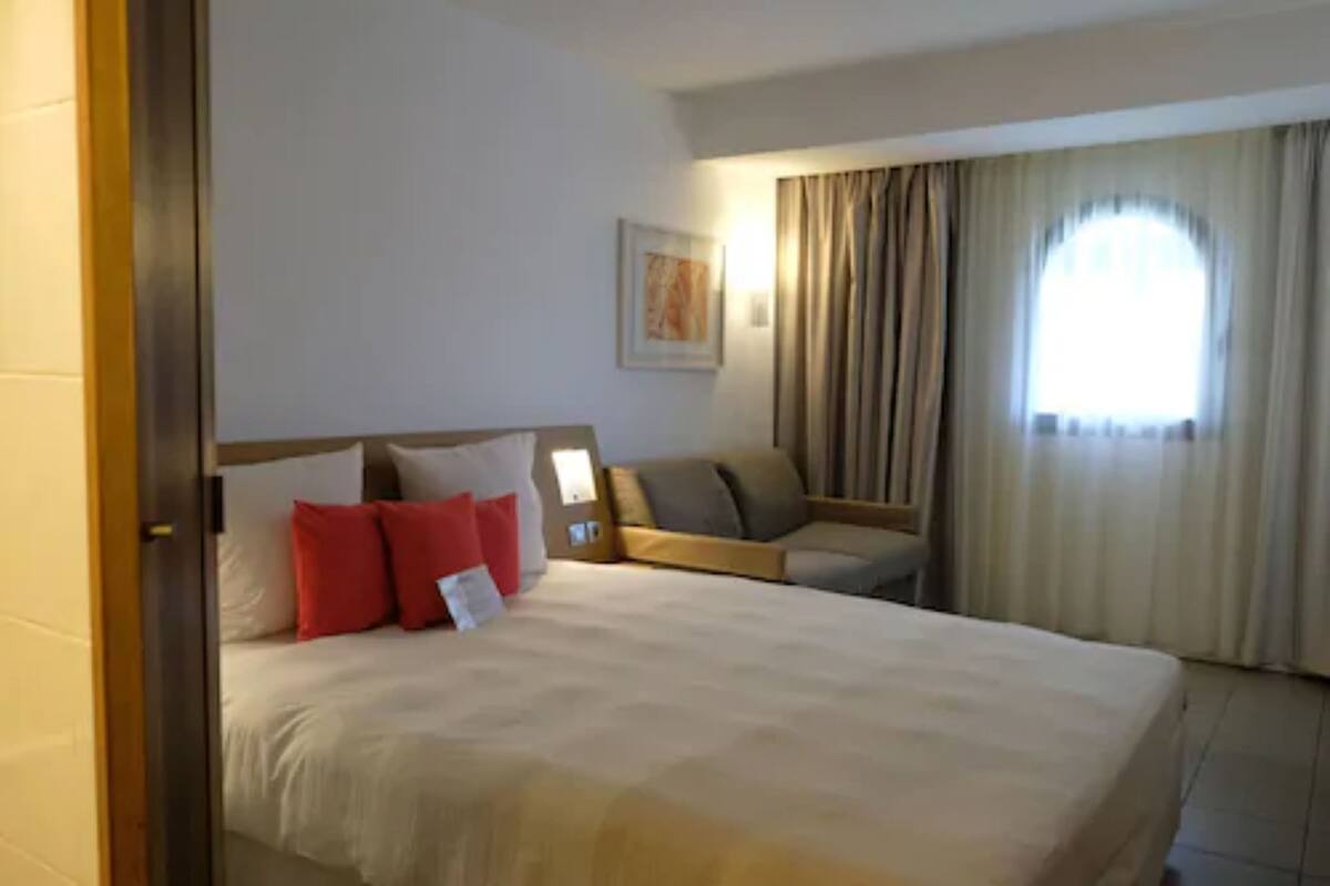 Une chambre d’hôtel avec un grand lit, un petit canapé, une fenêtre avec des rideaux et un tableau encadré au mur.