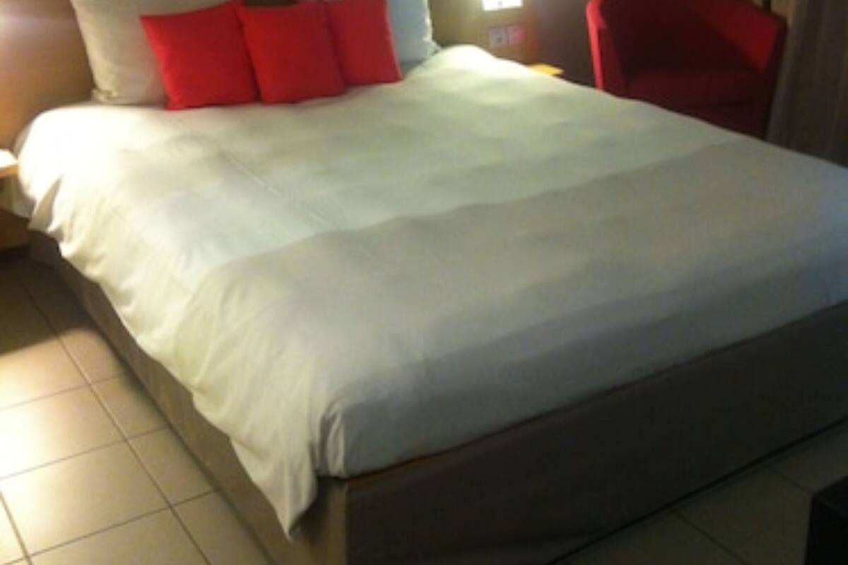 Une chambre d’hôtel avec un grand lit, une table de chevet, un fauteuil rouge et une lampe fixée au mur.