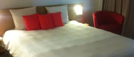 A hotel room with a large bed, a nightstand, a red chair, and a wall-mounted light.