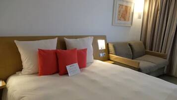 A hotel room with a bed, a nightstand, a reading light, a sofa, and a curtain.