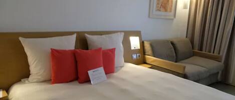A hotel room with a bed, a nightstand, a reading light, a sofa, and a curtain.