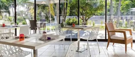 Un área de comedor al aire libre con sillas y mesas blancas, ventanas amplias y follaje en el fondo.