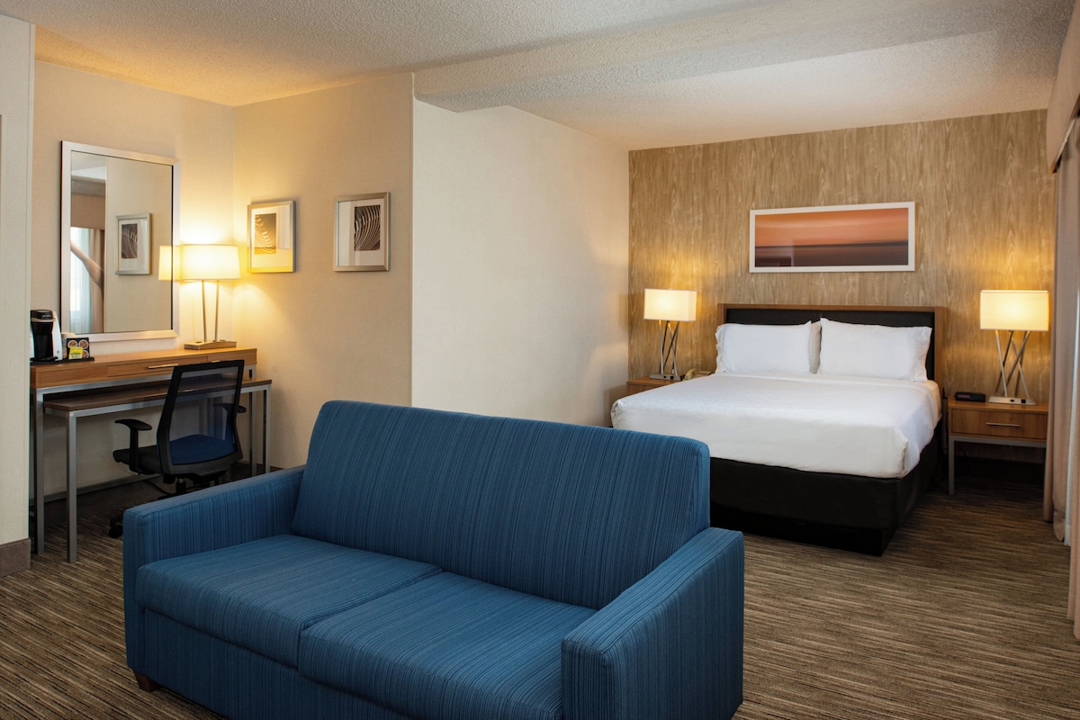 Quarto de hotel com cama, escrivaninha, poltrona e mesa de cabeceira com luminária.