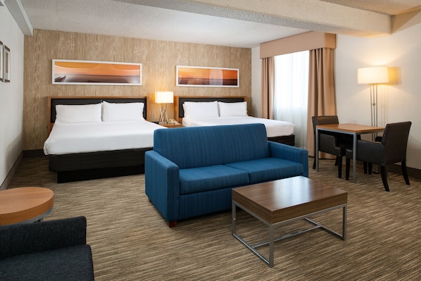 Quarto de hotel com duas camas, um sofá azul, uma mesa de centro de madeira, uma escrivaninha com duas cadeiras e duas luminárias de parede.