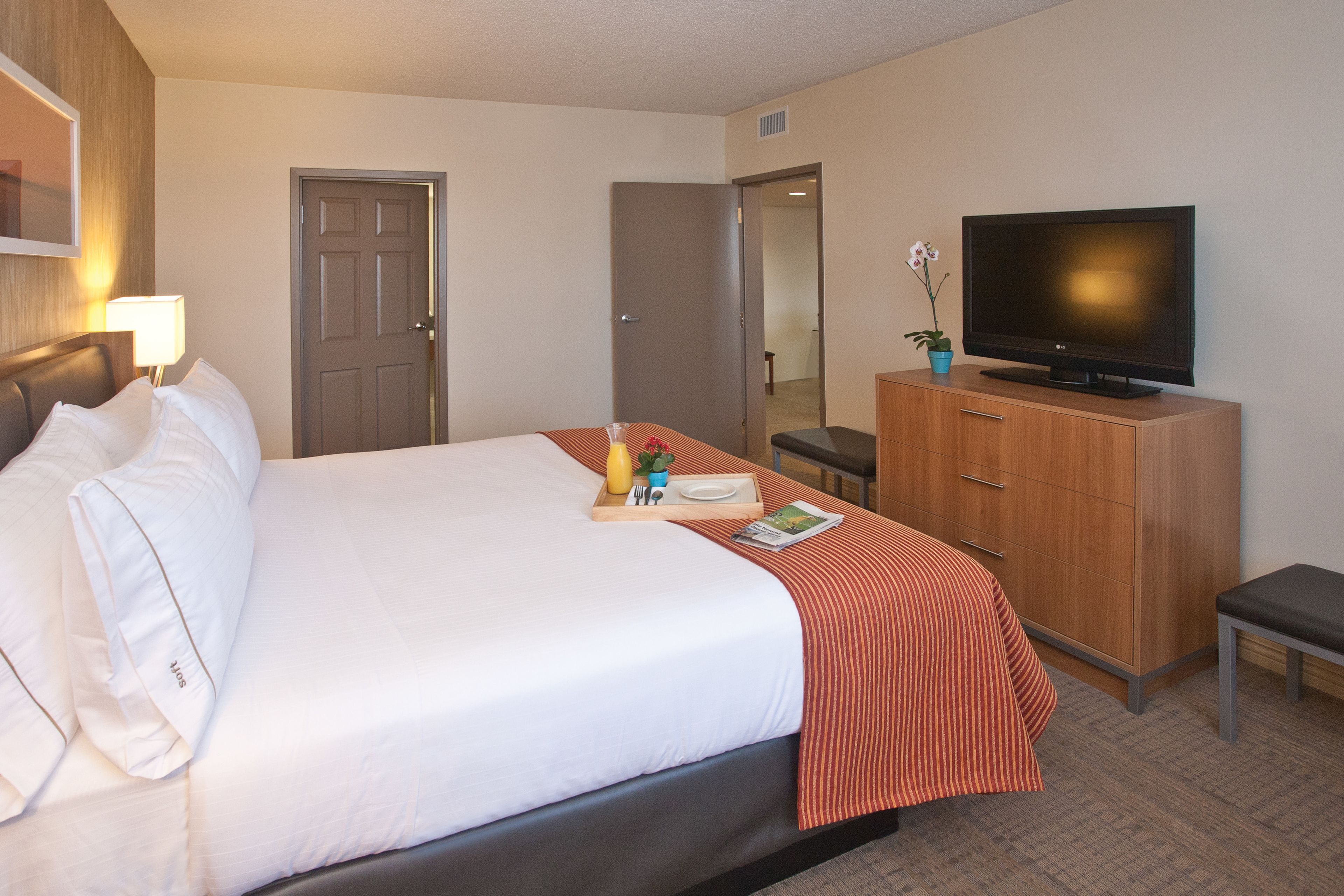 A hotel room with a bed, a television on a wooden cabinet, a desk with a chair, and a door leading to another room.