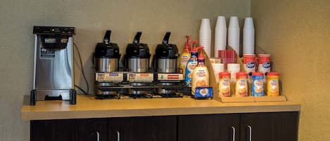 Una estación de café con una máquina de café, varios preparadores, vasos desechables y dispensadores de leche.