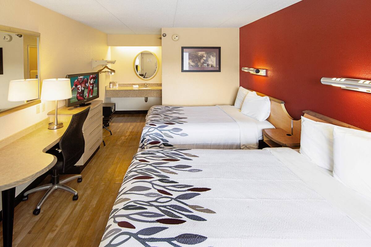 A hotel room with two beds, a desk, a chair, a TV, a mirror, and a bathroom.