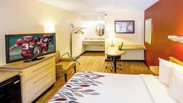 Quarto de hotel com uma cama, escrivaninha, cadeira, televisão, luminária de mesa, espelho e um quadro na parede.