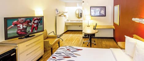 A hotel room with a bed, a desk, a chair, a television, a desk lamp, a mirror, and a framed picture on the wall.