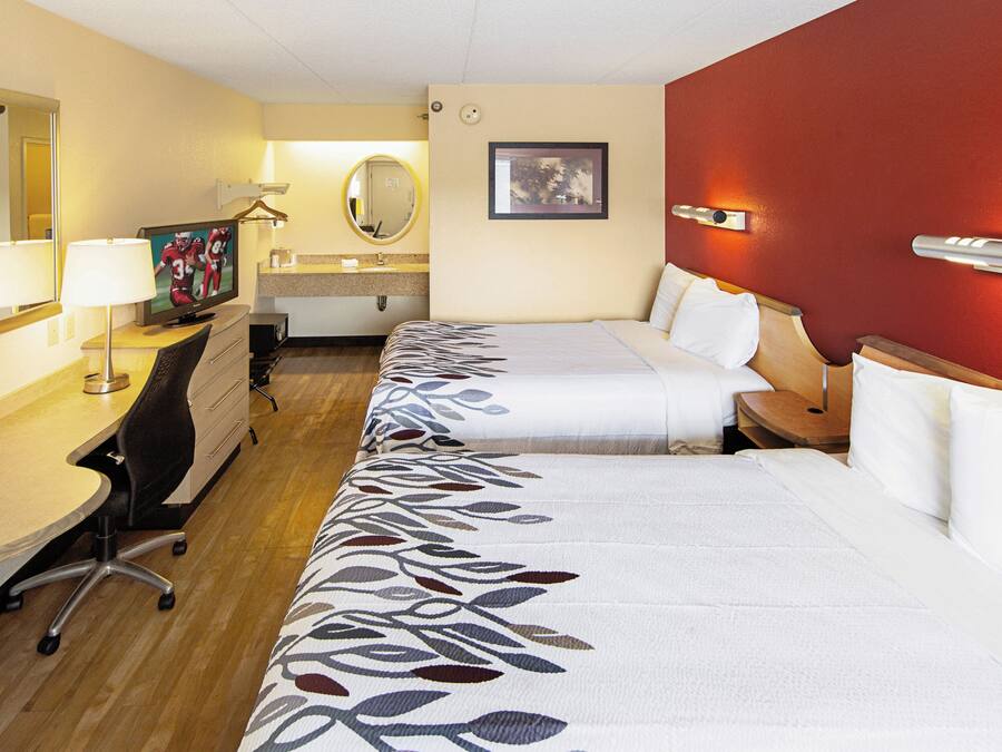 Quarto de hotel com duas camas, uma escrivaninha, uma cadeira, televisão, espelho e uma parede de destaque vermelha.
