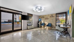 A modern hotel lobby with a large glass door, a seating area with chairs, a small desk, and a digital kiosk. - Days Inn by Wyndham Raleigh Midtown (Raleigh)
