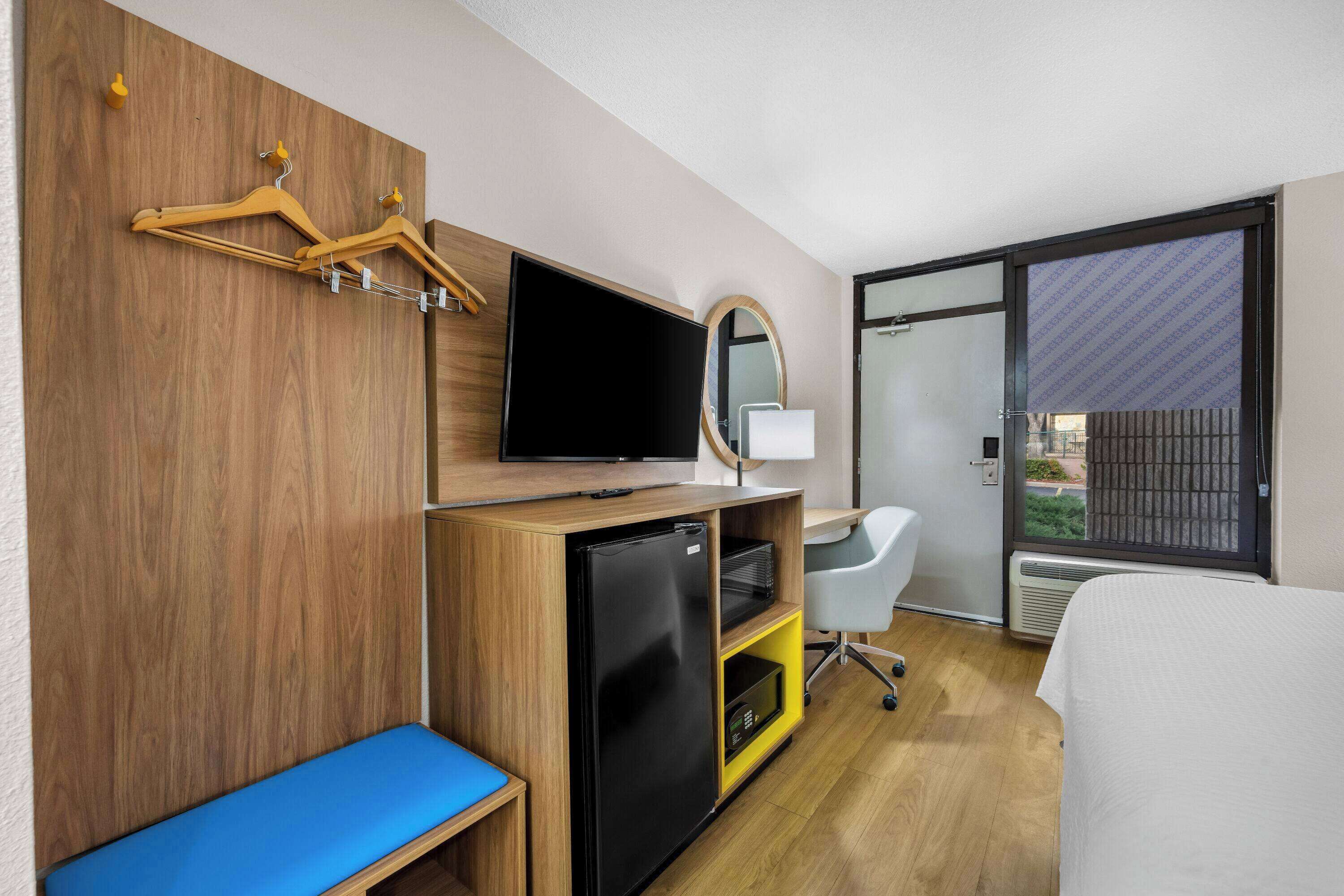 A hotel room with a wooden desk, a flat-screen TV, a small refrigerator, a blue bench, a white bed, and a large window with a view.