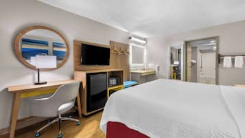 Habitación de hotel con una cama, un escritorio con silla, un televisor, un espejo y un baño visible a través de una puerta abierta.