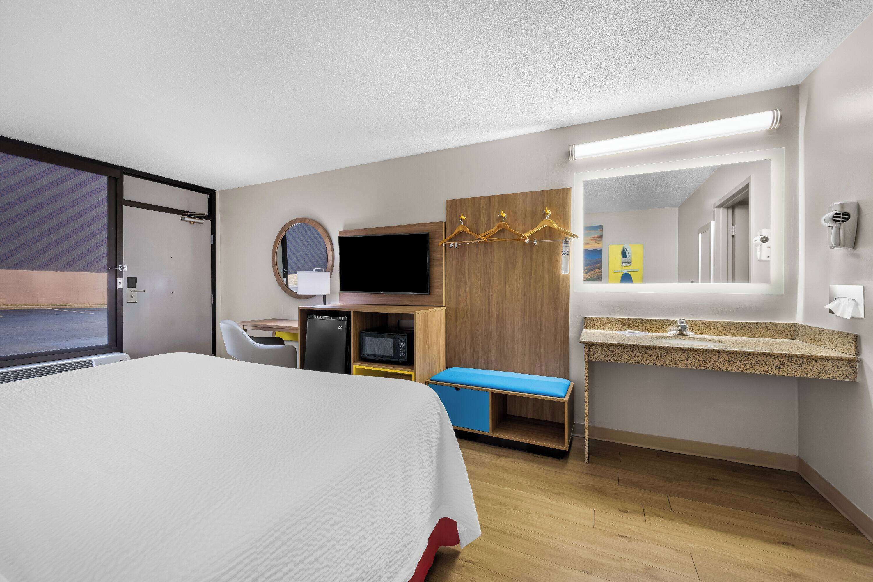 A hotel room with a bed, a desk with a computer, a TV, a microwave, a small seating area, and a bathroom with a sink and mirror.
