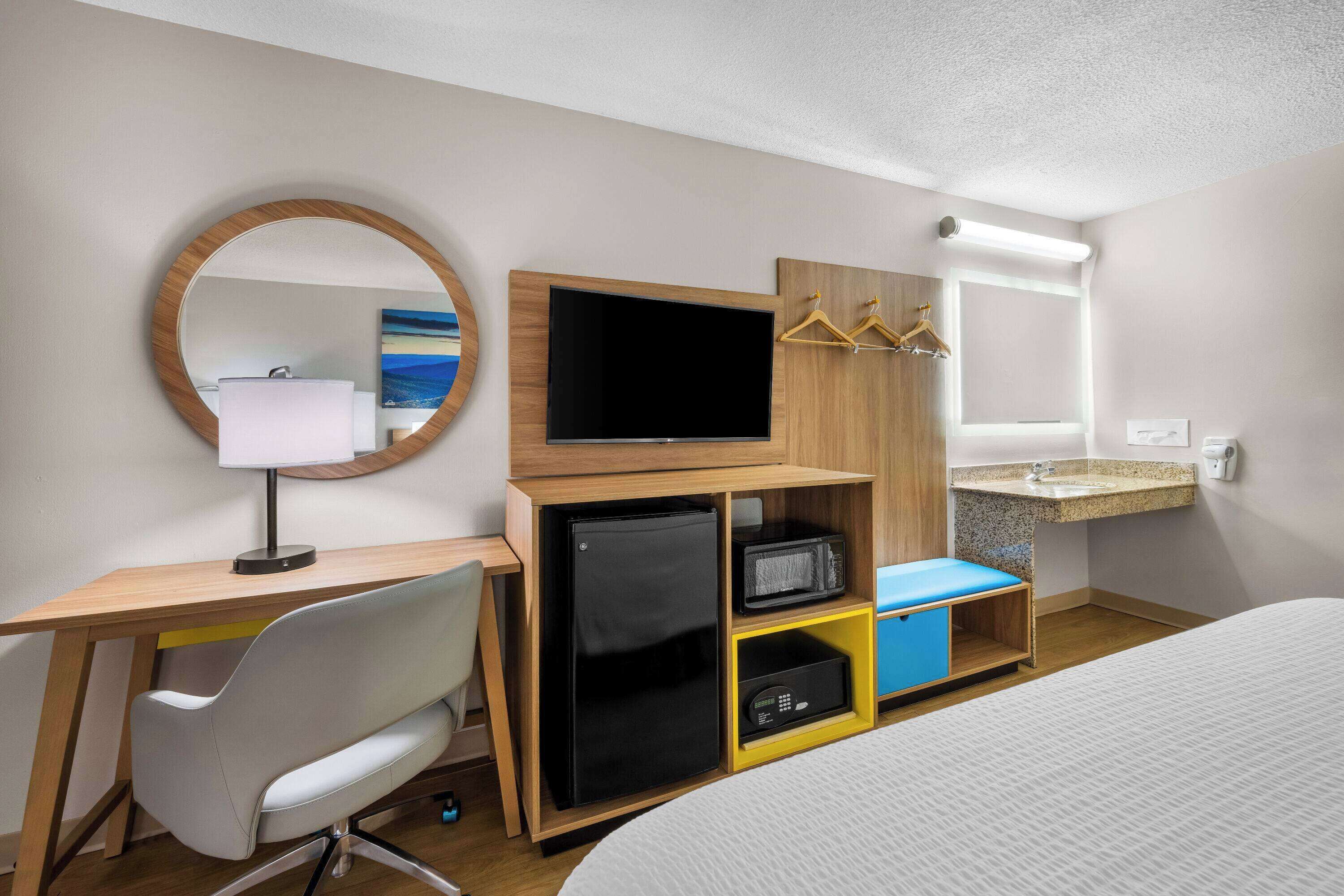 A hotel room with a bed, a desk with a computer, a TV, a mirror, a small fridge, a microwave, and a sink.