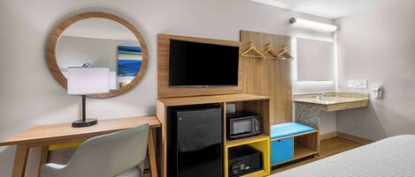 A hotel room with a bed, a desk with a computer, a TV, a mirror, a small fridge, a microwave, and a sink.