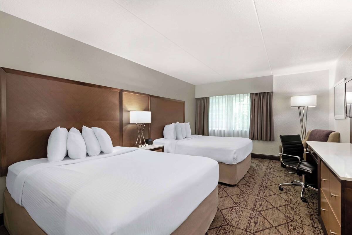 A hotel room with two beds, a desk, a chair, and a lamp.