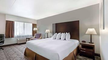 Quarto de hotel com uma cama grande, duas mesas de cabeceira com abajures, uma cadeira, uma escrivaninha e uma janela com cortinas.