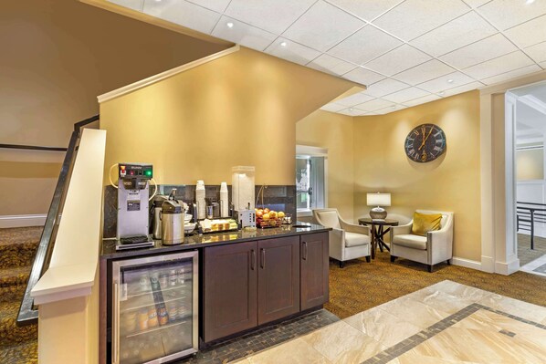 Vestíbulo de hotel con mostrador de recepción, máquina de café, refrigerador con bebidas, un reloj en la pared y un área de descanso con lámpara y sillón.