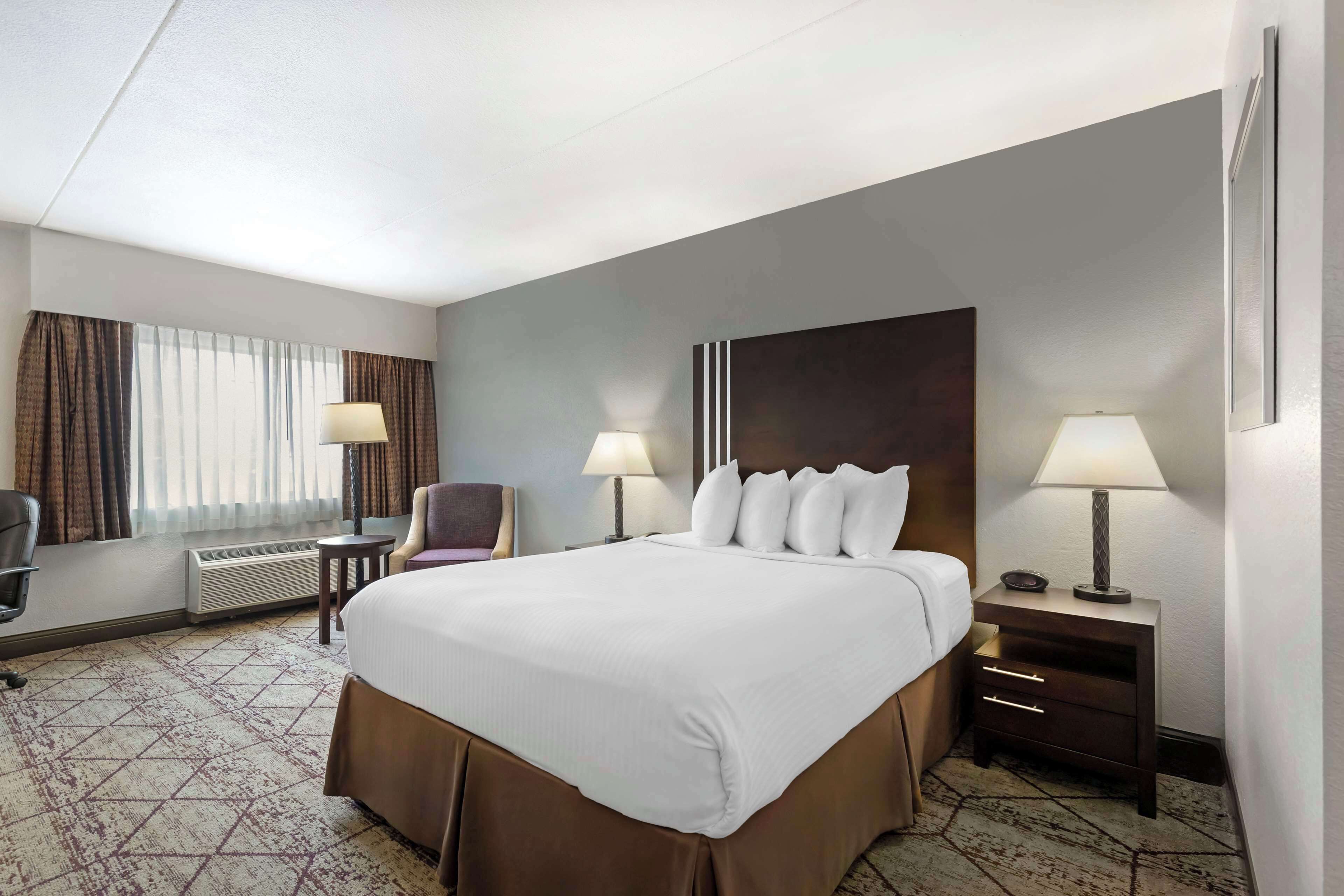 Una habitación de hotel con una cama grande, dos mesitas de noche con lámparas, una silla, un escritorio y una ventana con cortinas.