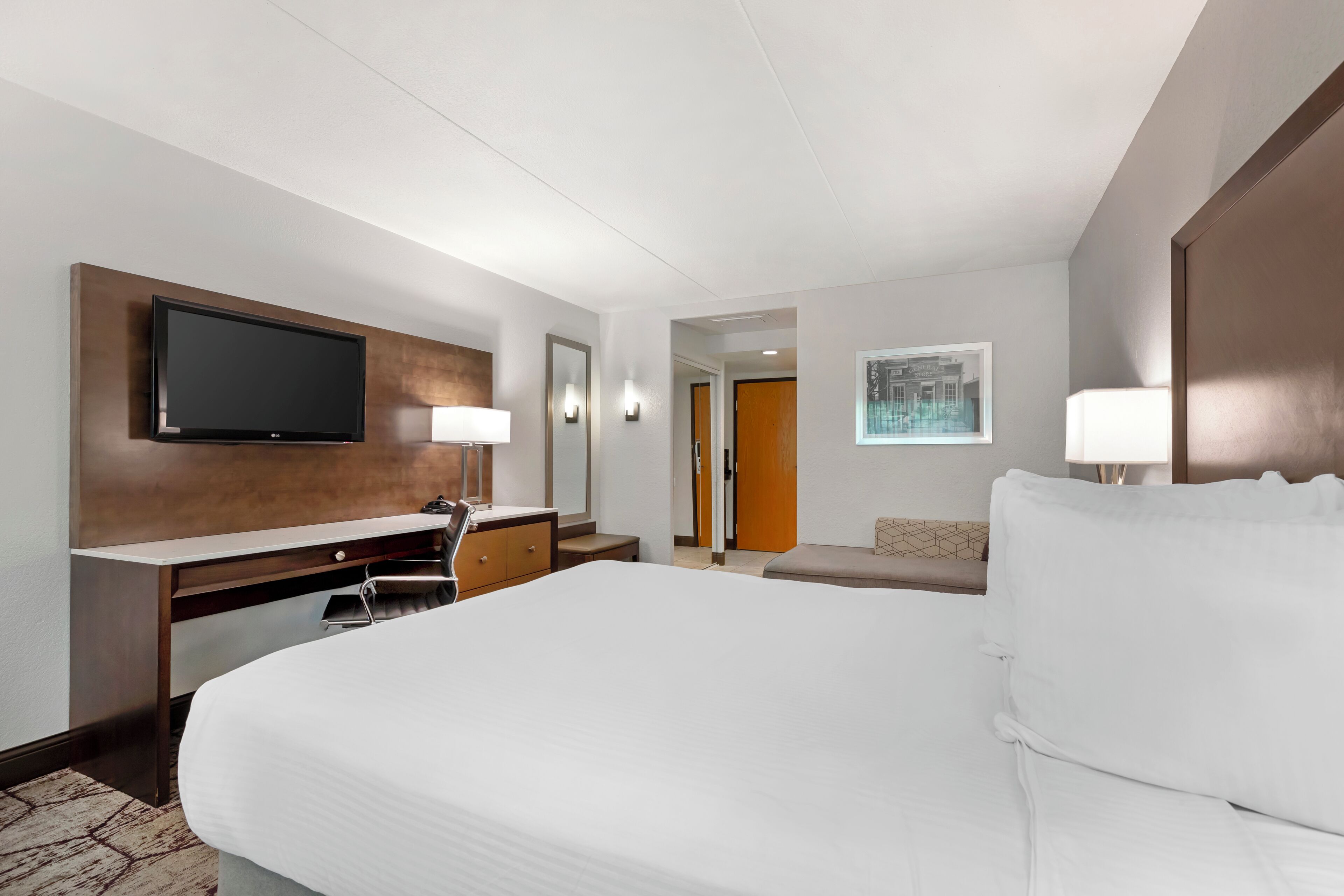 A hotel room with a large bed, a desk with a flat-screen TV, a chair, a lamp, and a painting on the wall.