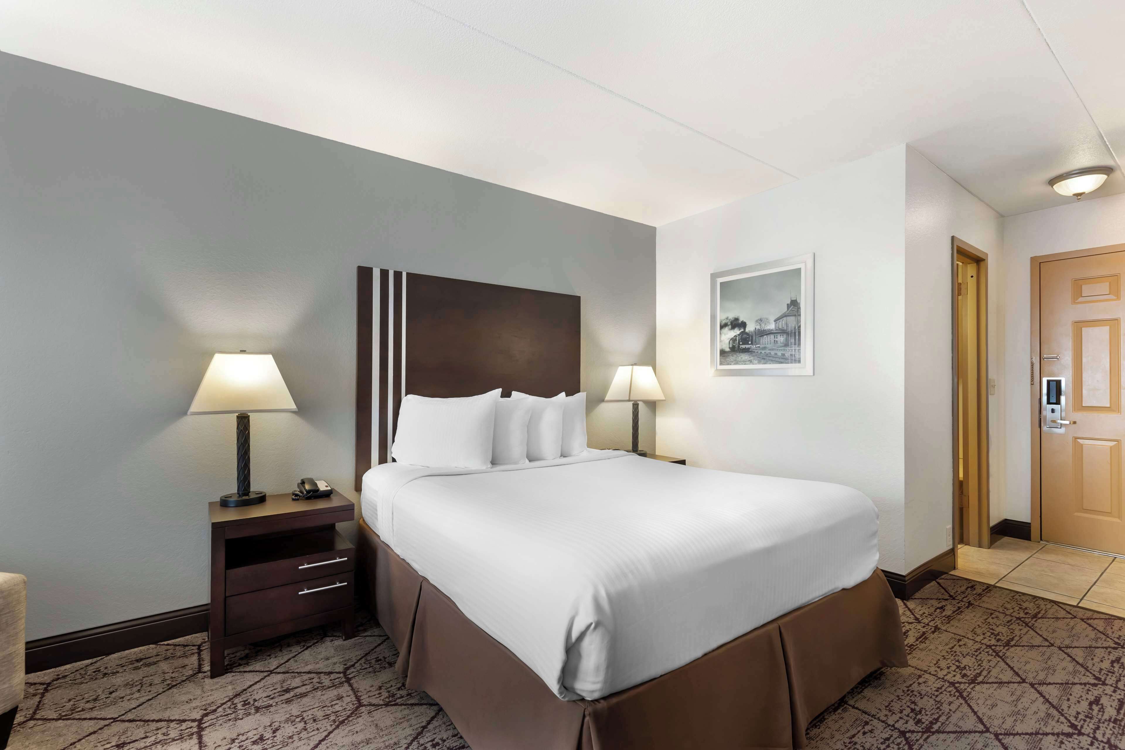 A hotel room with a large bed, two bedside lamps, a nightstand with a lamp, a framed picture on the wall, and a door with a keypad lock.