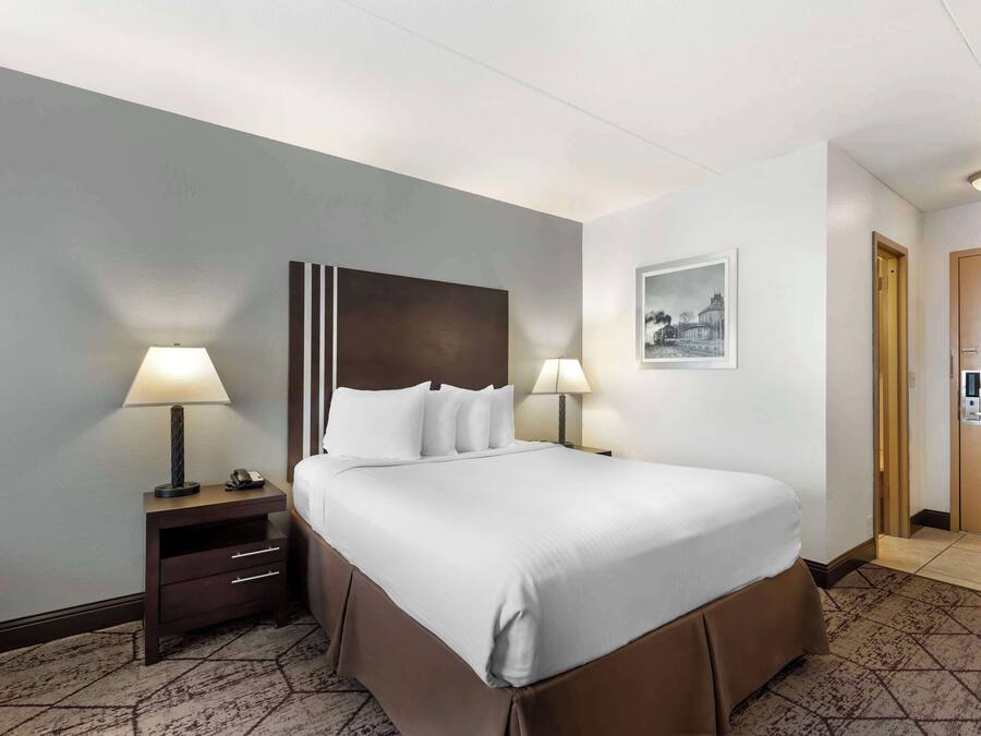 A hotel room with a large bed, two bedside lamps, a nightstand with a lamp, a framed picture on the wall, and a door with a keypad lock.