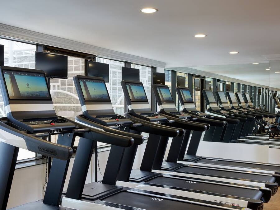 A row of treadmills with digital screens, a mirrored wall, and large windows.