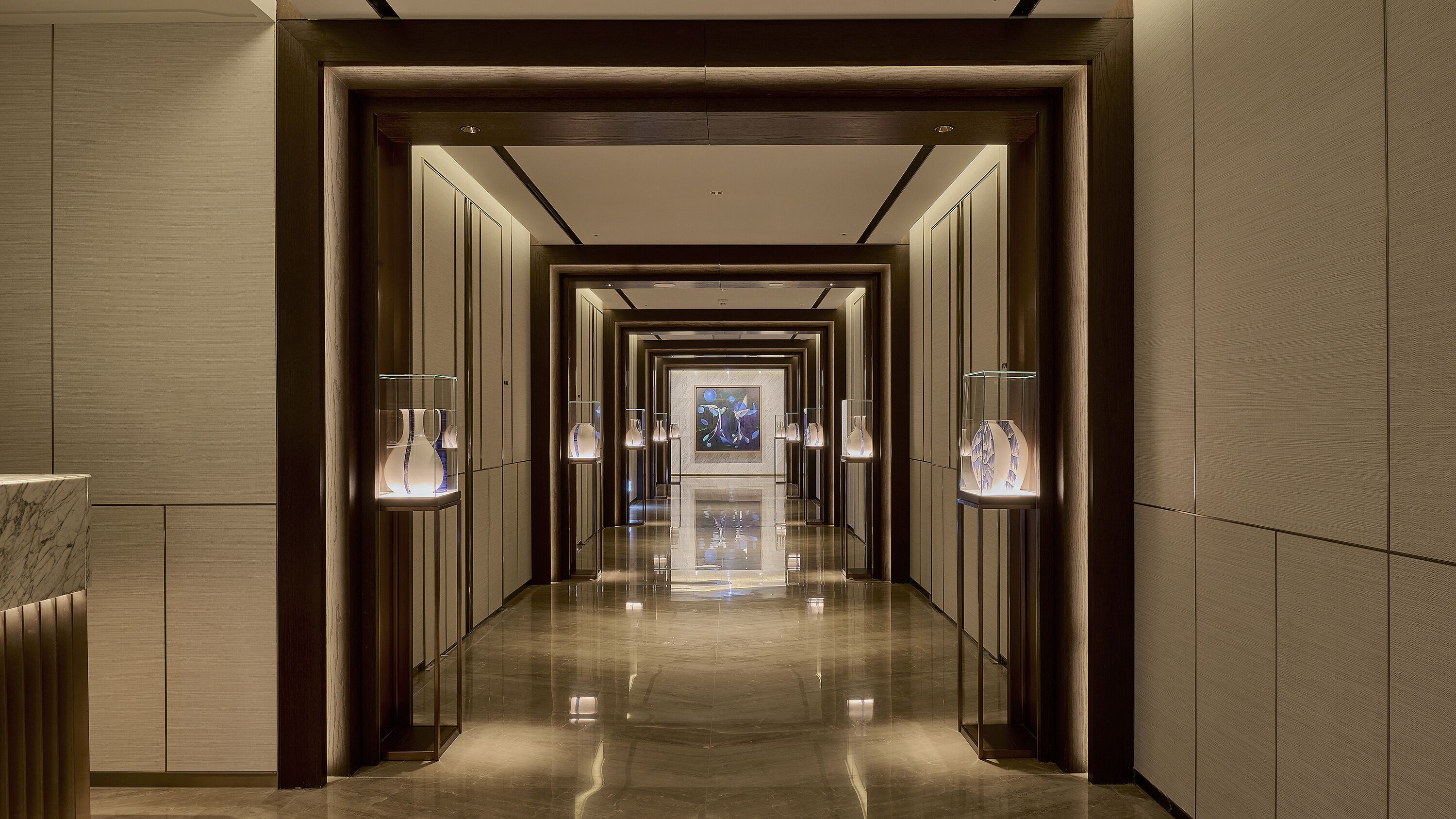 Un largo pasillo bien iluminado con piso pulido, flanqueado por vitrinas de vidrio con jarrones y una obra de arte central.