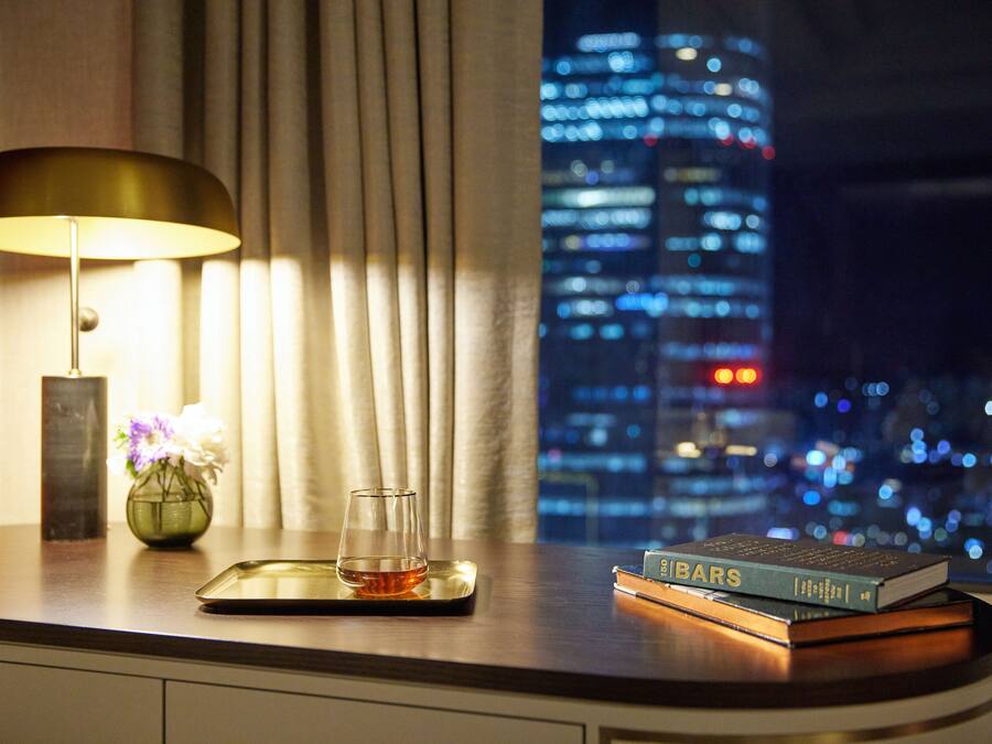 A night view of a cityscape through a window, a lamp, a vase with flowers, and a tray with a glass and a book on a table.