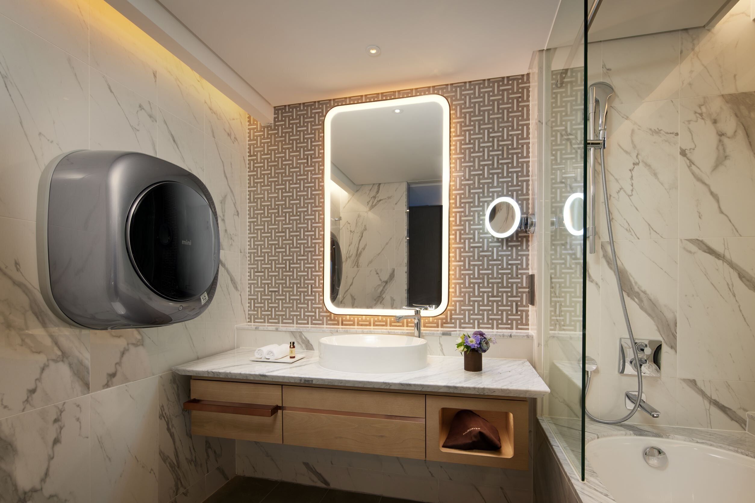 A modern bathroom with a marble sink, a handheld soap dispenser, a mirror with integrated lighting, a shower, and a patterned wall backsplash.