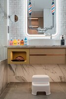 Un baño con espejo, lavamanos e inodoro. Hay un taburete pequeño frente al inodoro.