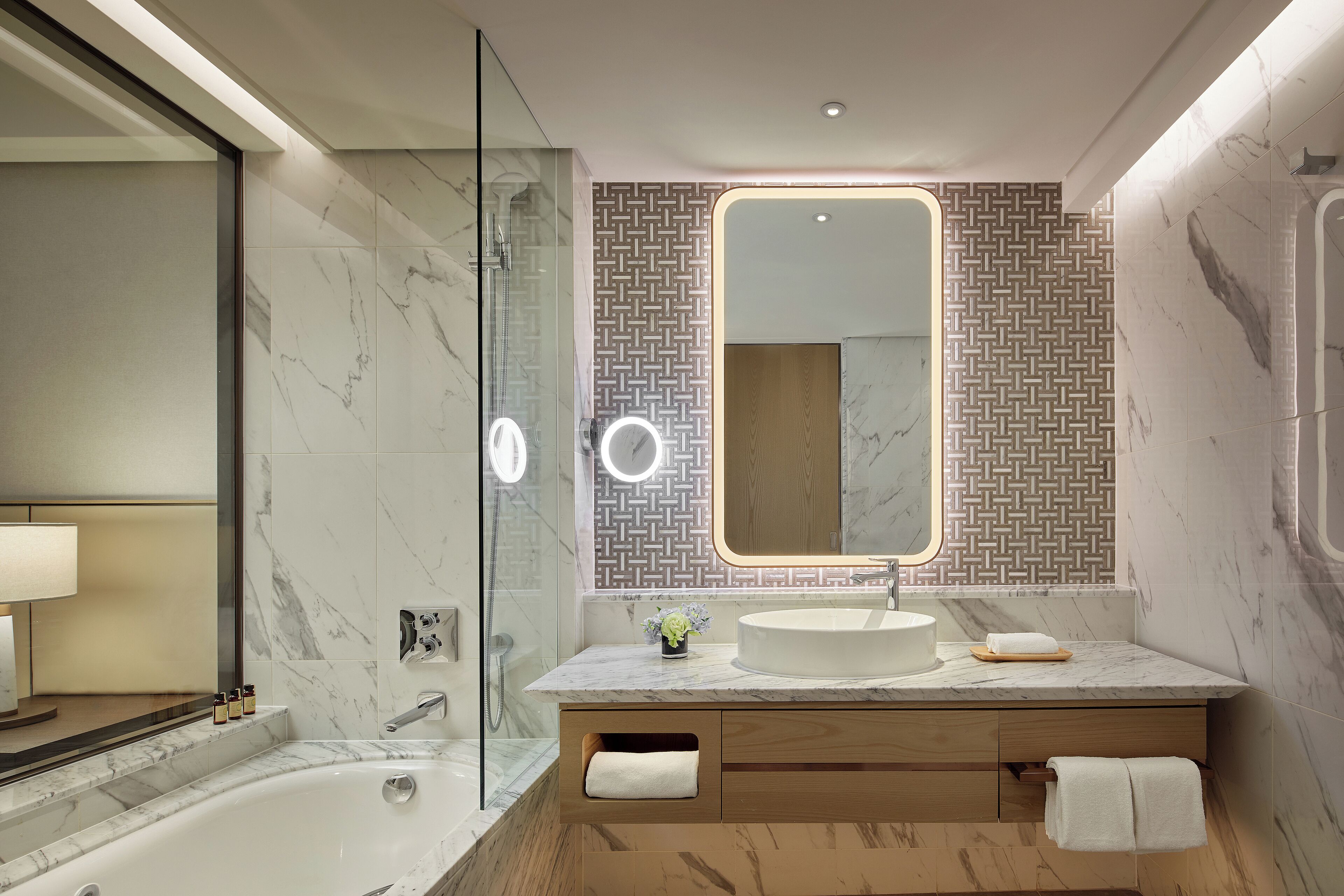 A modern bathroom with a glass-enclosed shower, a round mirror with integrated lighting, a marble sink, and wooden cabinetry.