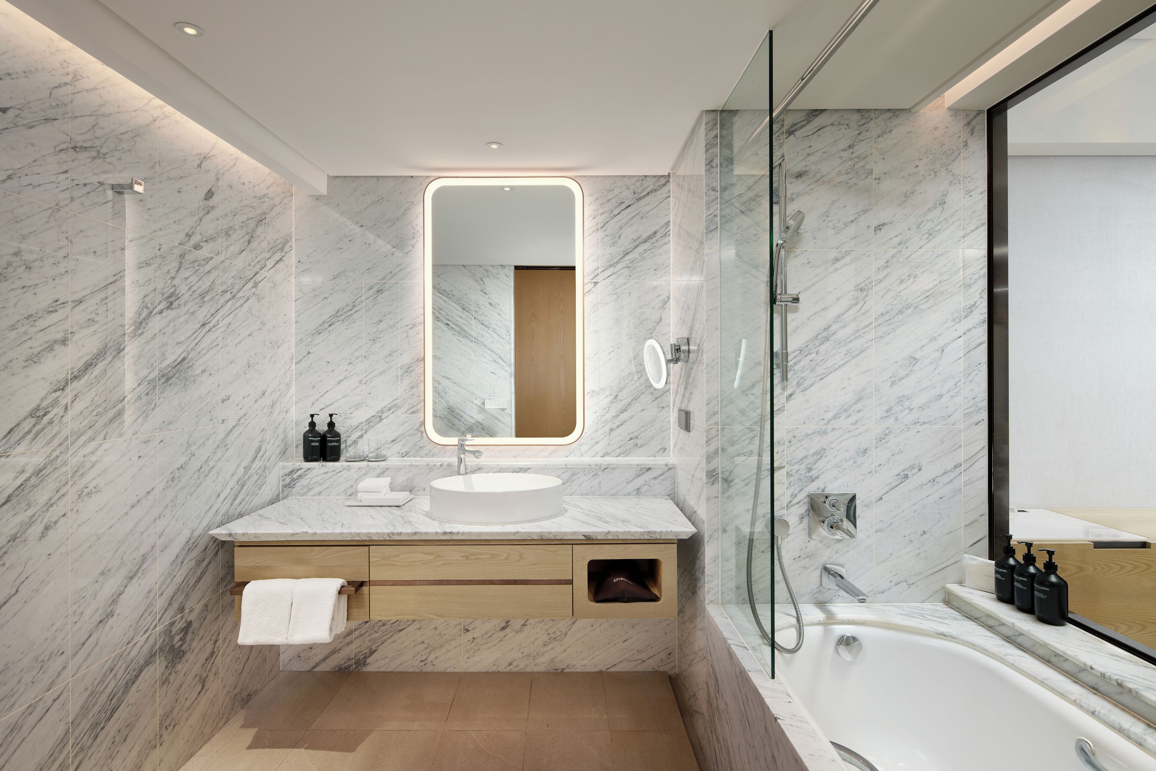A modern bathroom with a marble wall, a round mirror, a sink, a glass shower enclosure, and a bathtub.
