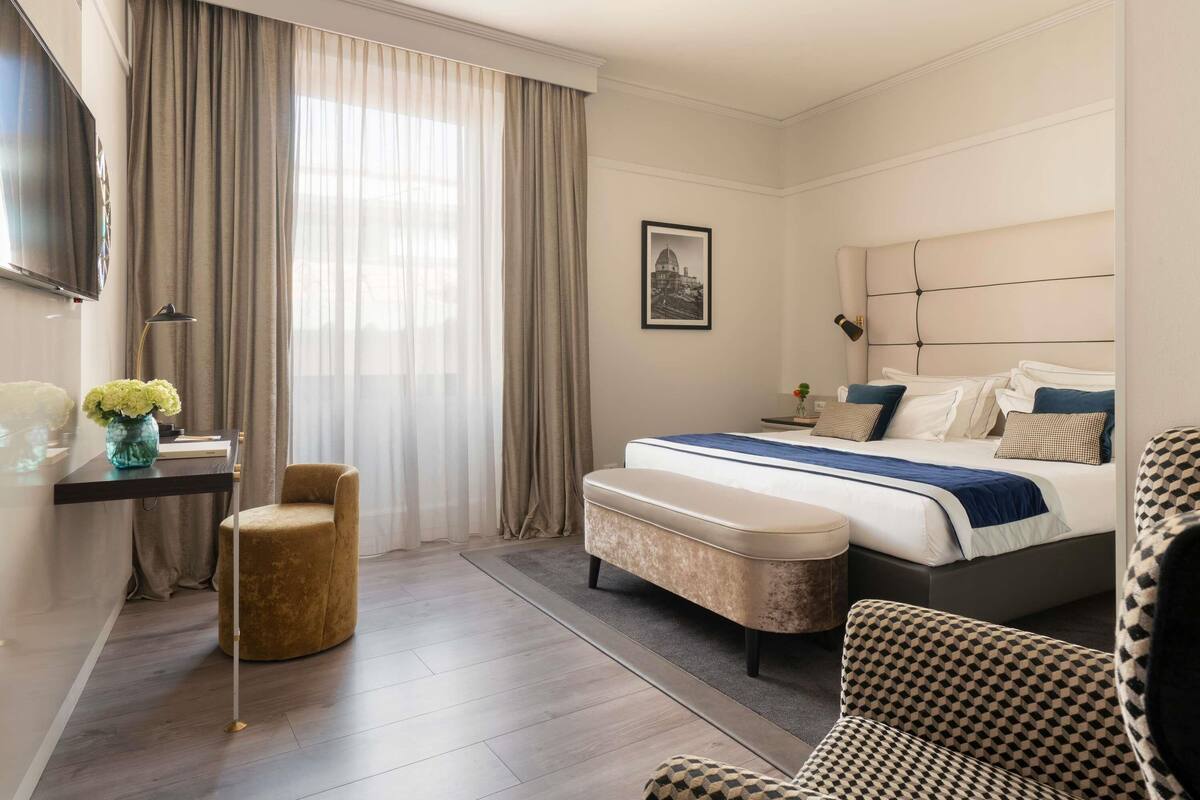 A modern hotel room with a large bed, a desk with a lamp, a chair, and a television on the wall.