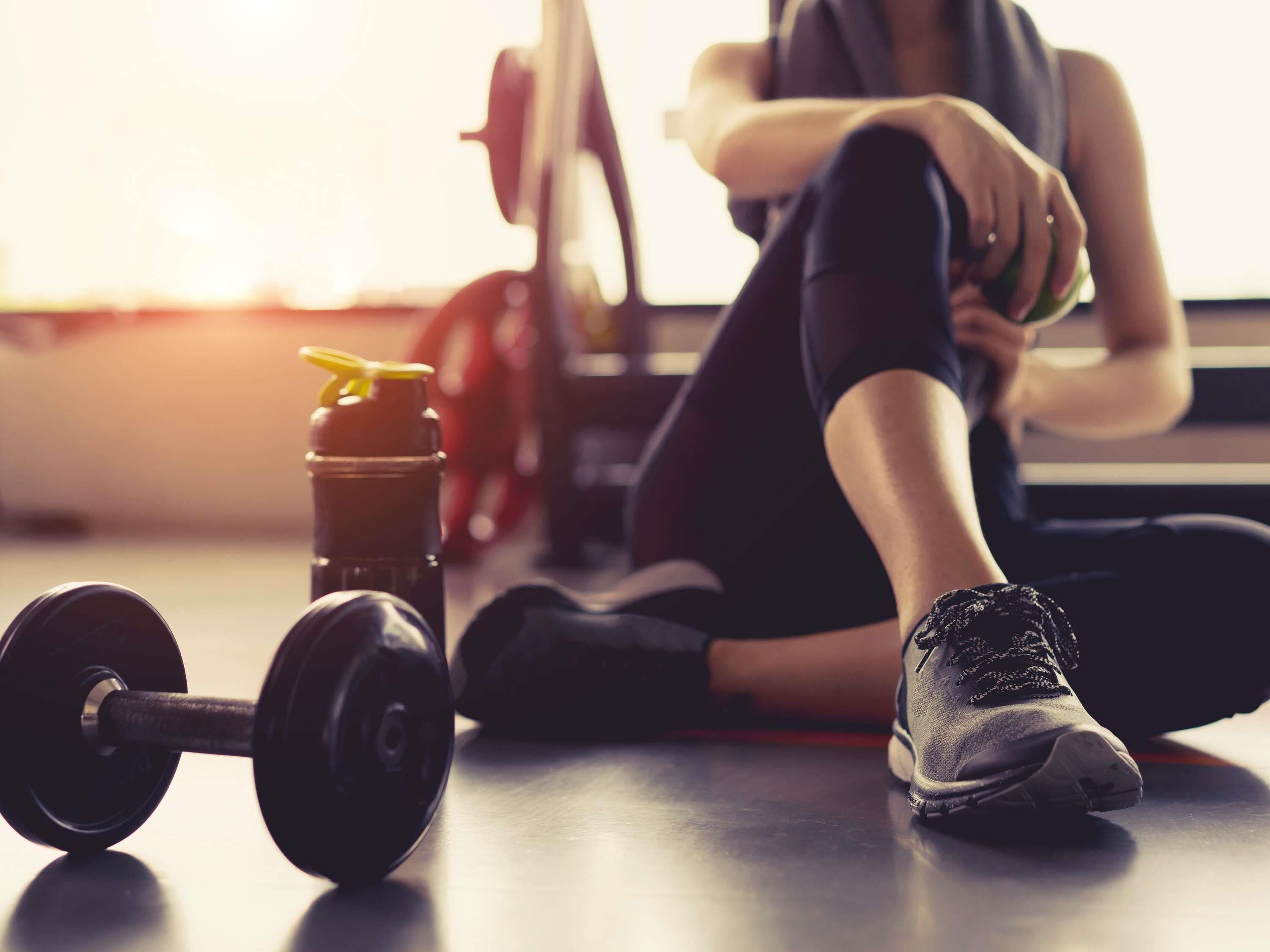 A gym setting with a dumbbell, a water bottle, and a person sitting on the floor.