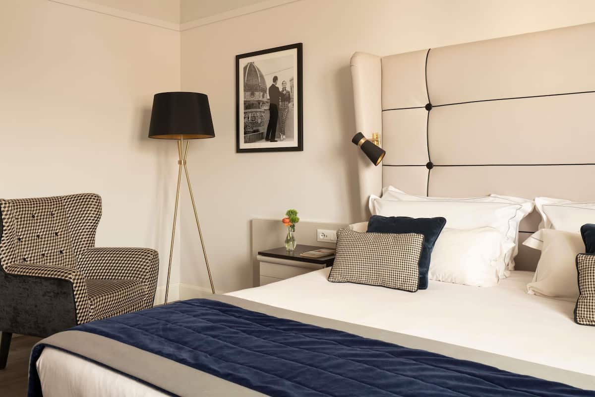 A hotel room with a large bed, a chair, a nightstand with a lamp, and a framed picture on the wall.