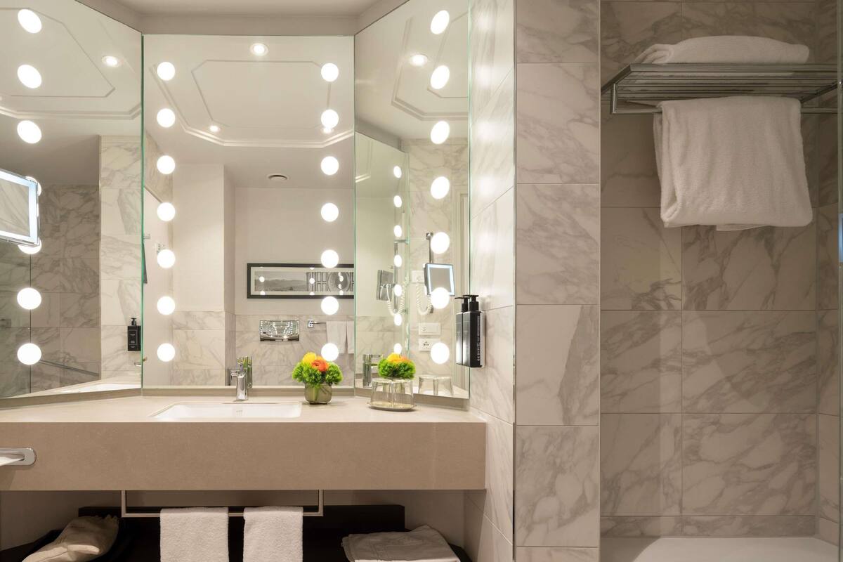 A modern bathroom with a vanity, large mirror with lights, marble tiles, and a towel rack.