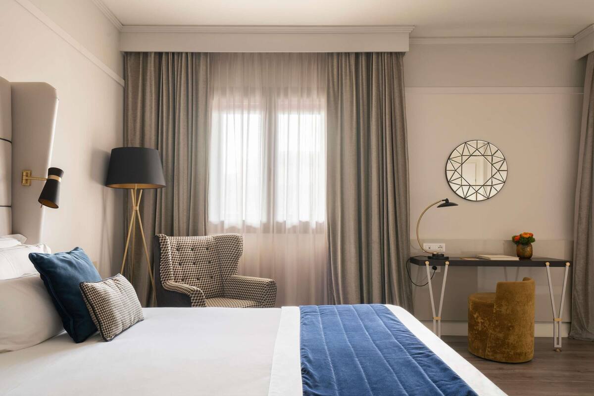 A hotel room with a bed, a desk, a chair, and a lamp.