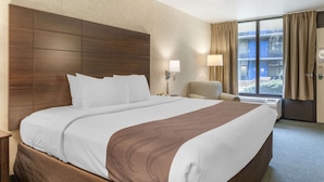 Una habitación de hotel con una cama grande, una mesita de noche, una silla, una ventana con cortinas y una lámpara de pared.