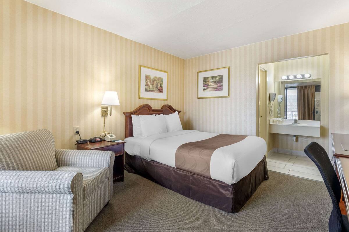 A hotel room with a bed, a chair, a desk, a lamp, a phone, and a mirror with lights.