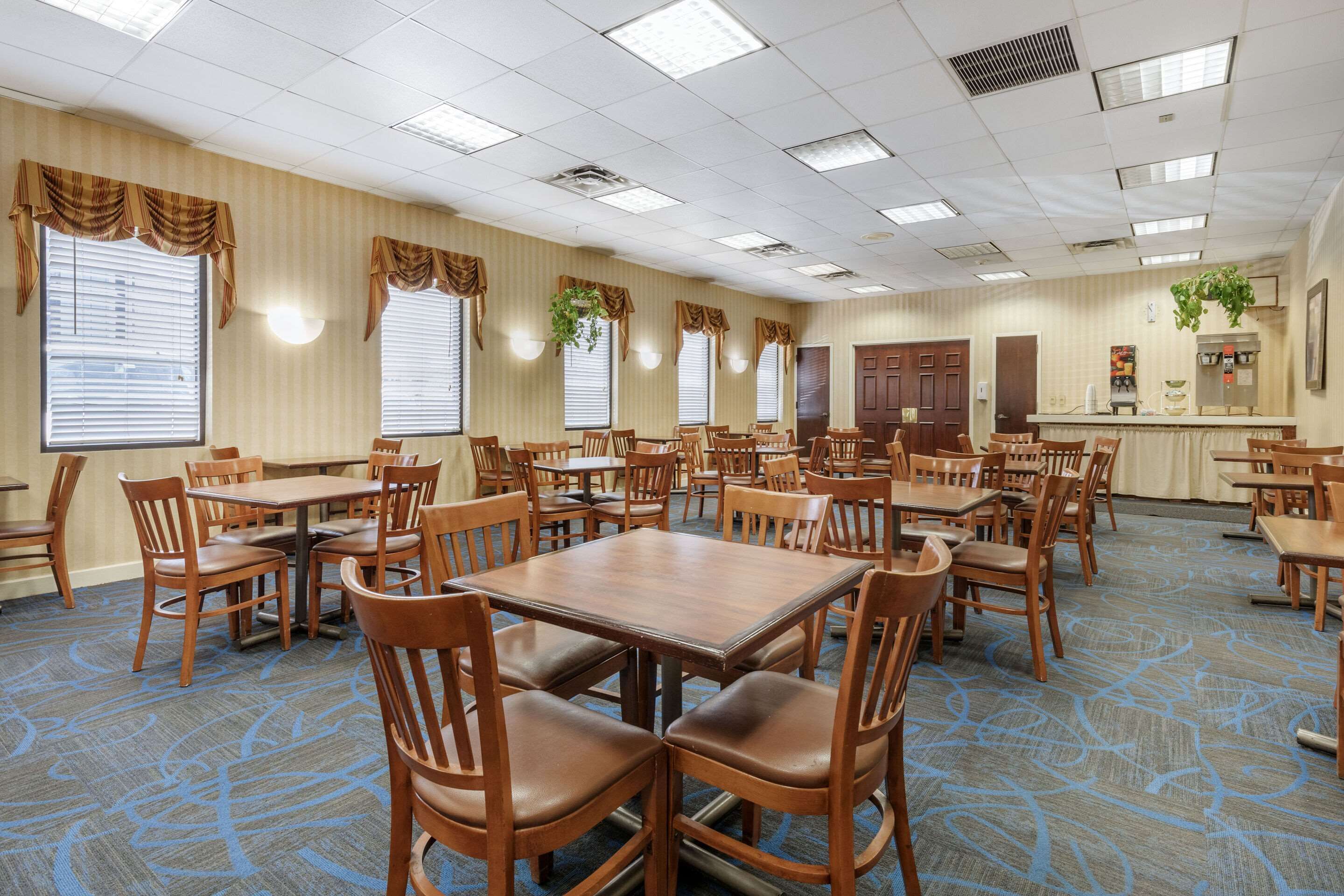 A hotel dining area with multiple tables and chairs, large windows with curtains, and a carpeted floor.