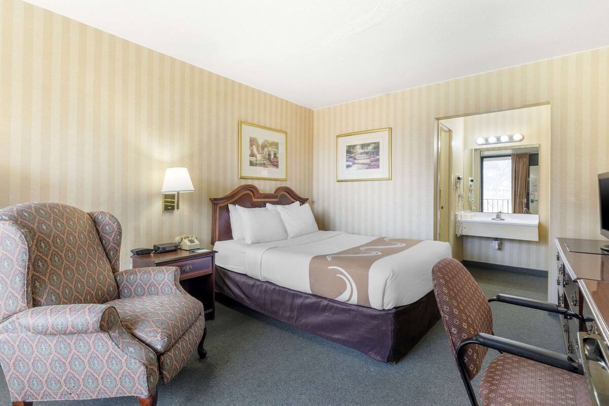 A hotel room with a bed, two armchairs, a desk, a mirror with lights, and framed pictures on the wall.