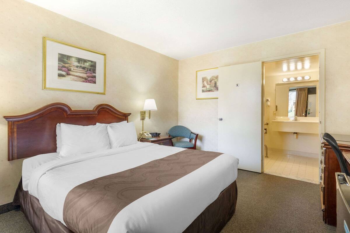 A hotel room with a large bed, a desk with a lamp, a chair, and a bathroom visible through an open door.