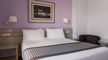 A hotel room with a neatly made bed, two wall-mounted lamps, a framed picture of a cityscape, and a small table with a chair.