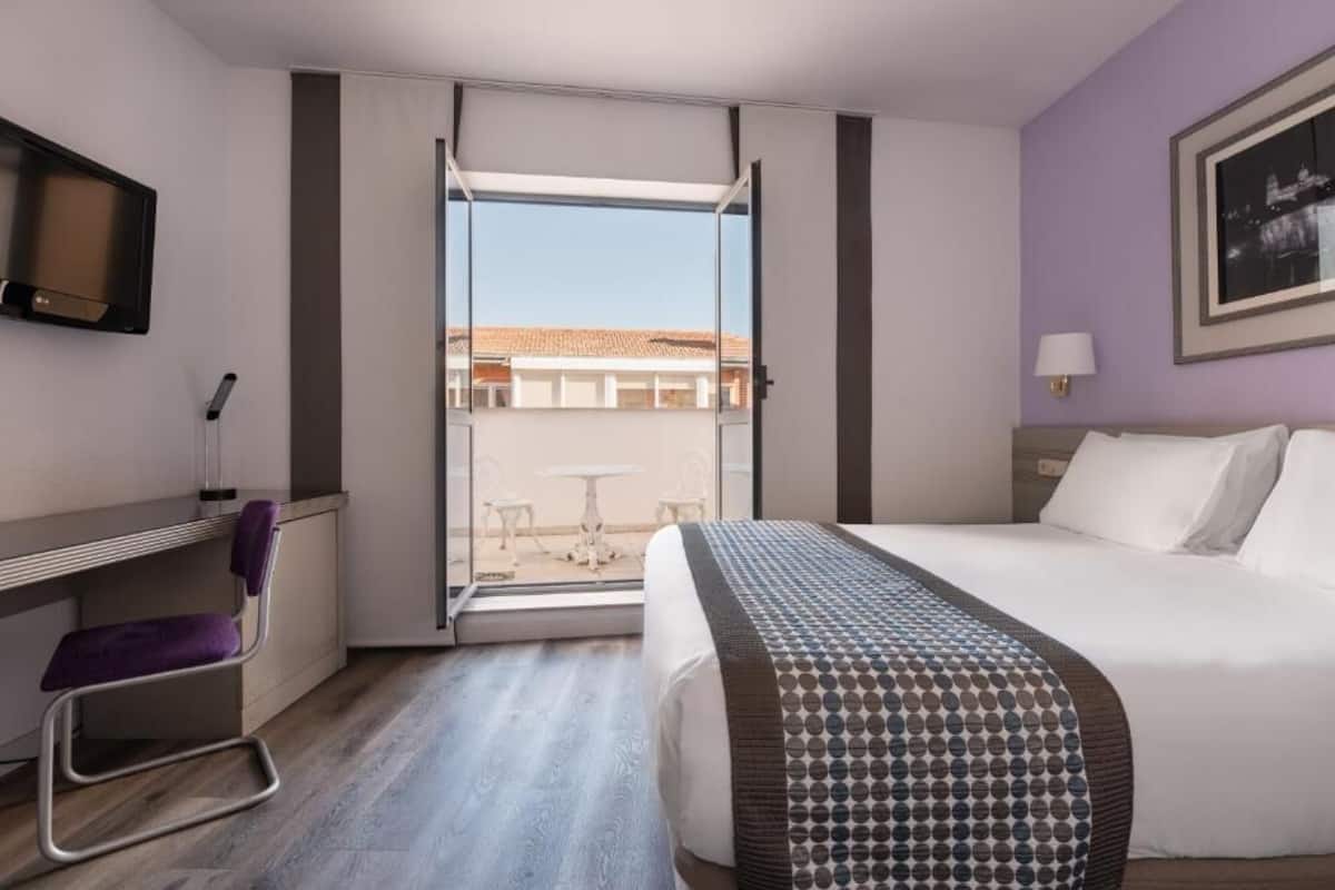 A hotel room with a bed, a desk, a chair, a TV, and a balcony with a view.