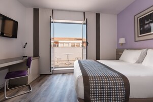 A hotel room with a bed, a desk, a chair, a TV, and a window with a view of buildings.